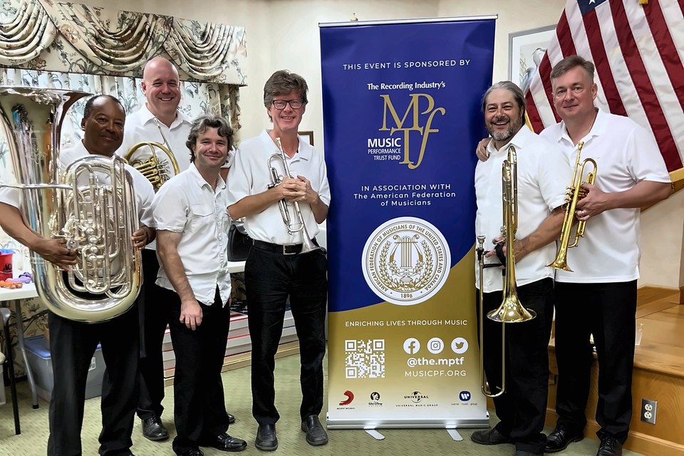 A brass group holding their instruments beside a Music Performance Trust Fund banner and an American flag